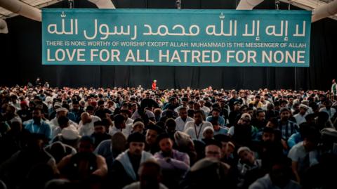 A crowd of people sat in a building under a large banner which says "There is none worthy of worship except Allah Muhammad is the messenger of Allah" "Love for all Hatred for none"