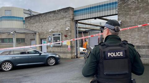 A police officer stands with his back to the camera and is stood near a grey, unmarked police car. There is a police cordon closing off the station and a white Audi car in the distance in the station.