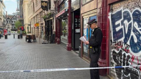 A police officer stands behind police tape on a city street in central London, reading notes outside a closed shop with graffiti on its shutter, while nearby streets are quiet and cordoned off.