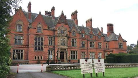 A Victorian red brick building with dozens of windows in front of a lawn. Two signs are wedged into the grass giving directions to the mansion and Wroxall Court. 