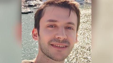 A young man with short brown hair smiles into the camera as he takes a selfie in front of a sparkling stretch of water with small boats in the top right corner.