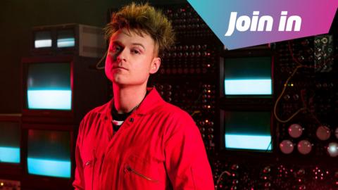 A white man has messy blonde/brown straight hair. He is stood in a big computer room with lots of wires and machines behind him. He is wearing a red boiler suit. In the right hand corner is a graphic that says 'join in' 
