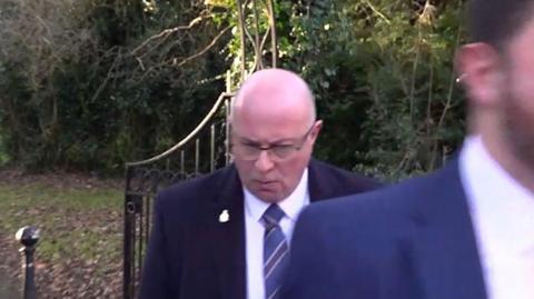 PC Mark Roberts leaving Durham Crown Court. He is wearing a dark suit and a blue striped tie. He is bald, is wearing glasses and looking down.