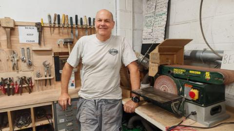 Mike Wallin stands in the Spelthorne Shed. He is wearing a grey t-shirt and grey work trousers. He leans against a work bench, with a circular saw to his left and several racks of woodworking tools to his right. He is a bald middle-aged man and is smiling.