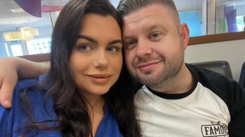 Josh Bashford wears a black and white t-shirt. He has short brown hair and a cropped beard. He hugs his wife Chloe who has long dark hair and wears a bright blue blouse. Both of them are smiling. 