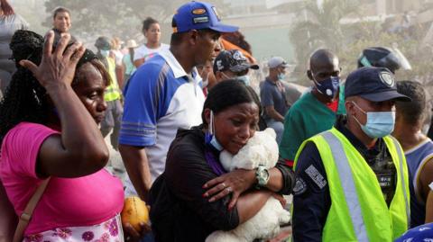 A woman in black with a face mask around her chin cuddles a white teddy bear for comfort, looking distressed. She stands among a crowd of others, including men in masks, scrub tops, high-vis yellow vests. A woman to her right in a pink and floral dress has a hand on her head.