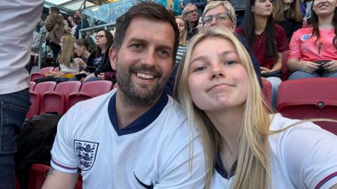 Richard and Imogen wearing England football tops sitting in the stands at a football stadium taking a selfie. They are both smiling at the camera. She has long blonde hair which is straight while he has dark hair and a short beard.