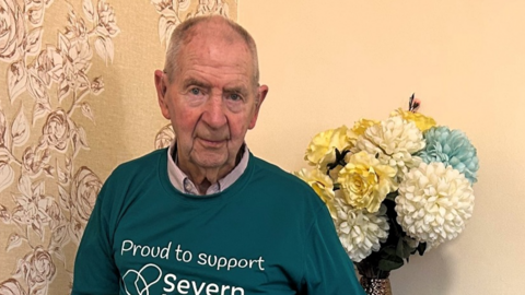 A man with short grey hair and a teal jumper a room with a large bunch of white flowers behind him