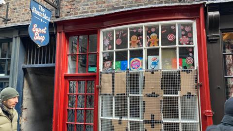 The Shambles Sweet Shop in York - a red facade set in an old brick building. Six window panes are boarded up with cardboard and masking tape, with a white grill placed over the top.