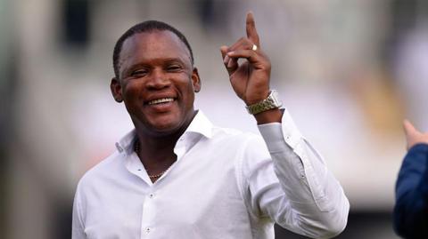 An upper body picture of Devon Malcolm who is wearing a white shirt and has one arm raised in the air. He is smiling. The background is blurred.