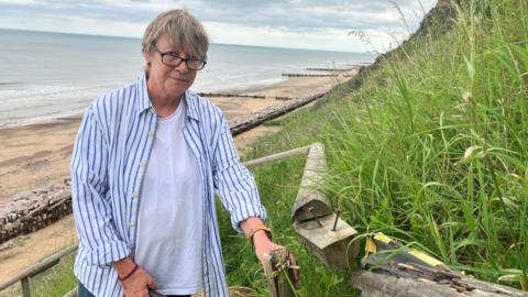 A woman wearing a striped shirt and glasses looks towards the camera while standing on a series of steps that lead down to a beach, next to handrails that have been damaged.