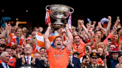 A man holding a trophy above his head, he is wearing an orange GAA top with crowds of people cheering behind him