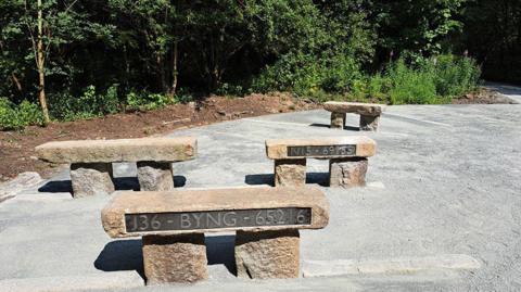 Stone benches which have metal plaques with different carvings are position on a walking trail in Carlisle. The benches, which are place in front of woodland, have a Roman-era design.