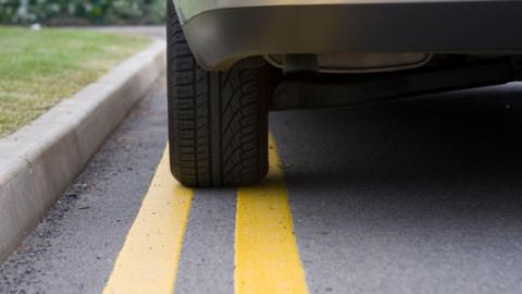 A stock image of a car parked on a double yellow line. Only the rear passenger side tyre and a small section of the rear of the car can be seen.