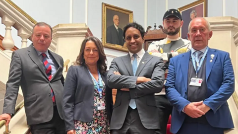 Five people stood on a grand staircase. Four are in business attire and one man is wearing casual clothes. They are Arron Banks, Linden Kemkaran, Zia Yusuf, Nathaniel Fried and Brian Collins.

