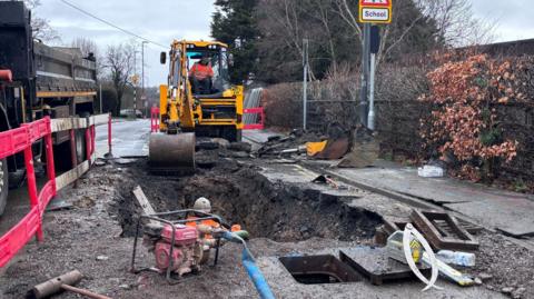 Crews work on a large hole in the street in Bristol as they repair a water main. In the background is a yellow mechanical digger