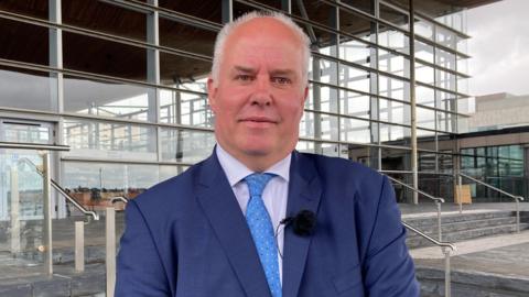 Andrew RT Davies wearing a dark blue suit, a light blue tie, a slightly purple shirt and a microphone stood on the steps in front of the Senedd building in Cardiff Bay.
