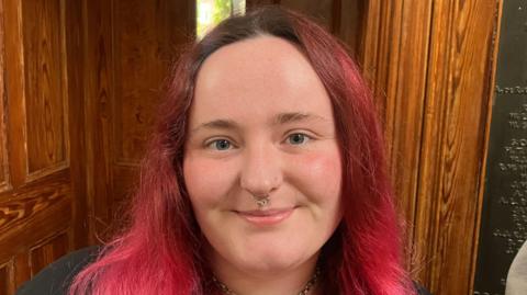 Ciara Kennedy has long dyed pink hair and blue eyes. She is standing in front of a wooden door smiling. She is wearing a silver choker necklace and finer silver chain below.