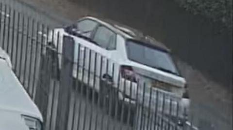 A grainy CCTV image of a white skoda driving along a road beside a metal barrier