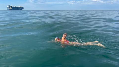 A picture of Melanie Barratt swimming in the blue sea in a colourful swimsuit and cap. There is a ship in the background.