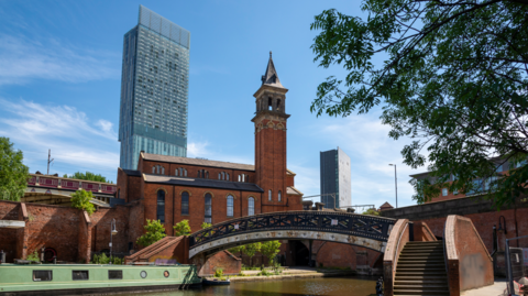 A shot of Manchester with a canal, bridge and boat in the foreground and the Beetham Tower in the background