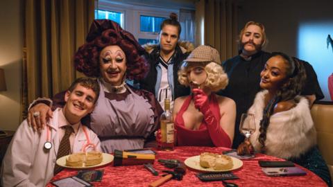 A group for people sit at a table in fancy dress. 

From Left to Right, Elijah Young as Stewart, Mark Benton as Mam, Charlotte Riley as Danni, Phil Dunning as Dickie, Patsy Lowe as Sal and Alexandra Mardell as Lucinda. 