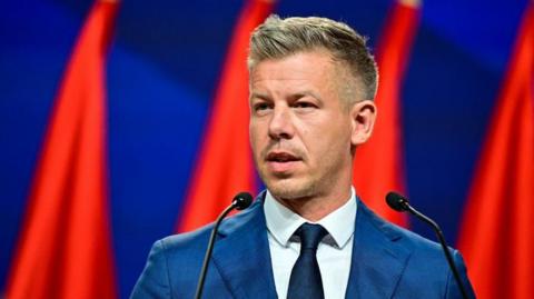 Magyar speaking into microphones with flags behind him. He is wearing a navy blue suit.