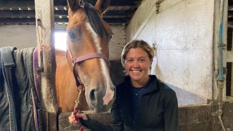 A smiling woman with blond hair and wearing a black fleece holding the reins of brown horse which is standing behind a wooden stable door