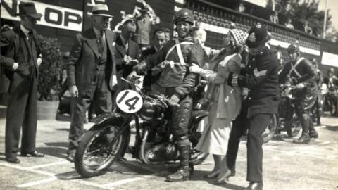 A black and white shot from the film showing George Formby as George Shuttleworth on his number 14 motorcycle at the TT grandstand surrounded by people.