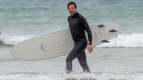 James Norton is walking out of the surf wearing a black wetsuit and carrying a surfboard.