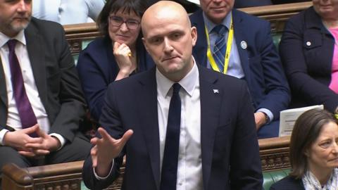 Stephen Flynn speaking from the green SNP benches in the House of Commons. He has a bald head and is wearing a blue suit jacket, a dark tie and a white shirt. He is gesticulating with his hand and there are other politicians around him. 