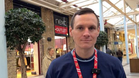 Sean Taylor stands in the atrium of the Swindon Outlet Centre while wearing a red lanyard. A potted tree and a number of shop fronts, including Next, can be seen behind him.