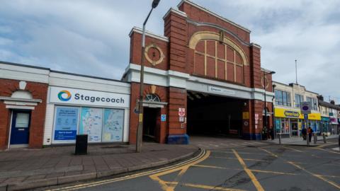 The outside of Workington bus station. It is a red-bricked building flanked by shops. 