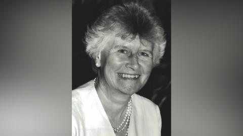 A black and white photograph of a smiling older woman, looking at the camera, with a pearl necklace and light blouse.