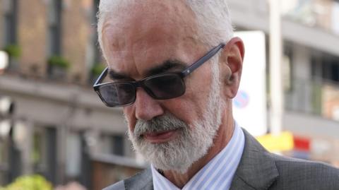 A close up of the face of Cdr Julian Bennett, wearing sunglasses - he has a grey beard and grey hair and he is looking down. 