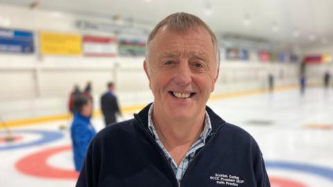 Keith Prentice, man in 70s with fair hair smiling, pictured with ice rink in background.