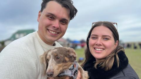 Kieran Smith, a man with dark hair and is wearing a cream coloured jumper. He is holding a brown and white dog which is wearing a grey harness. He is stood next to Megan Jones, a woman with mid length brown hair and blonde streaks who has a pair of sunglasses on her head and is wearing a black jacket.