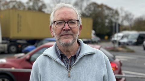A wide shot of Peter Dowd standing in front of a busy road. He has short white hair and is wearing a pair of glasses, a grey jumper, and a stripey shirt underneath.