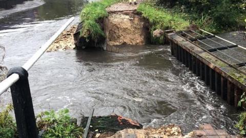 A photo taken from one side of a small weir looking towards a footpath on the other side of the bank