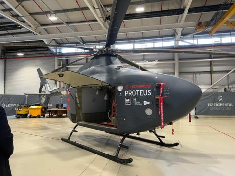 A helicopter has the word Proteus on the side. It has no windows. Its side door is open, and it is stationary in a hangar.