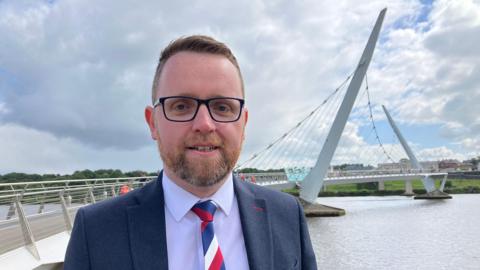 Gary Middleton wearing navy suit, white shirt and red, white and blue tie, pictured in front of the Foyle Peace Bridge.