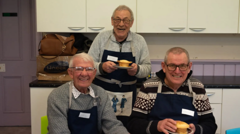 Three smiling older men in a kitchen wear aprons and hold two pies on plates