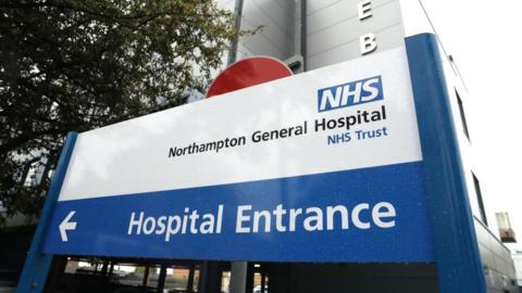 A blue and white sign. The top of the sign is white with the writing Northampton general hospital. The bottom of the sign is blue with a white arrow pointing to the left and writing saying Hospital Entrance.