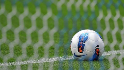 A picture of a ball that is behind a football net. It is white, blue and orange in colour. It is on the goaline which is a green grass field and white markings.