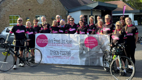About 20 cyclists wearing mainly black outfits stand with bikes and hold up a large mainly white banner including the words Worcestershire Breast Unit Haven. Everybody knows somebody are among other words printed on the banner. 