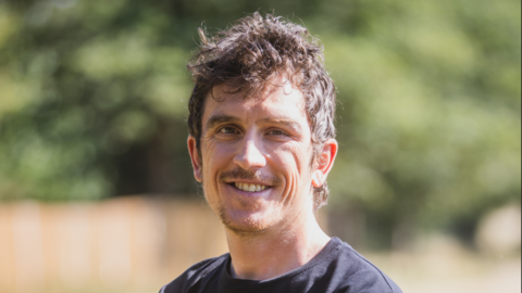 Geraint Thomas is in the middle of the image looking towards the camera smiling. He has short brown hair and a short moustache and beard and is wearing a black T-shirt. 