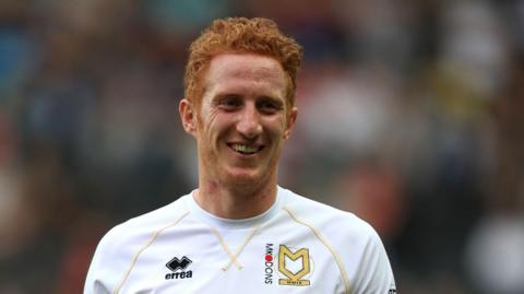 Dean Lewington is on a football pitch walking towards the camera. He has short ginger hair and a short bead. He is wearing a white football shirt with the MK Dons insignia on it.