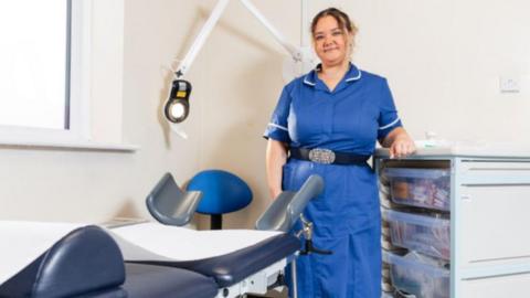 A woman in a blue work uniform standing next to trust equipment is looking at the camera.