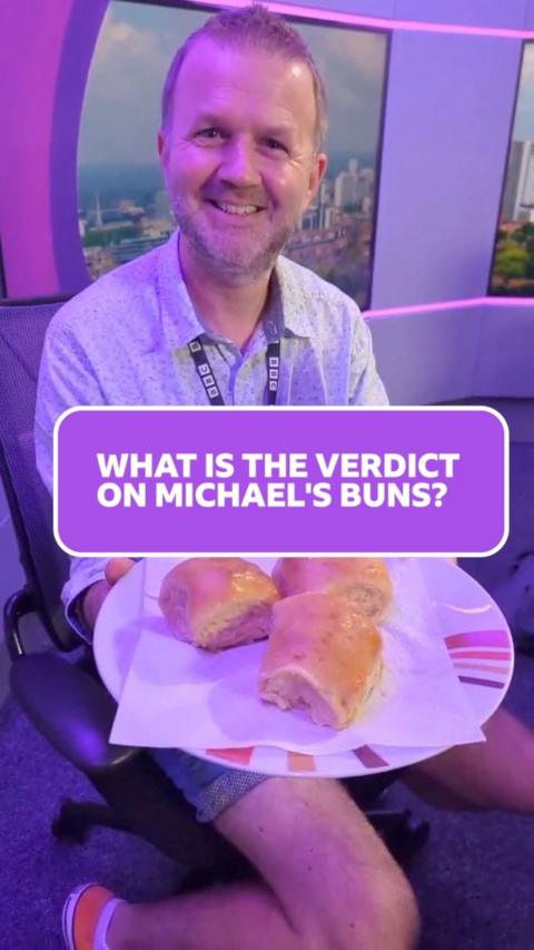 Newsreader Michael Coombes has short light brown hair and wears a shirt, with denim shorts and is holding a plate with three bread rolls towards the camera