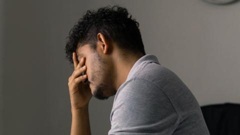 A man holds his head in his right hand with tiredness. He is wearing a grey tshirt.